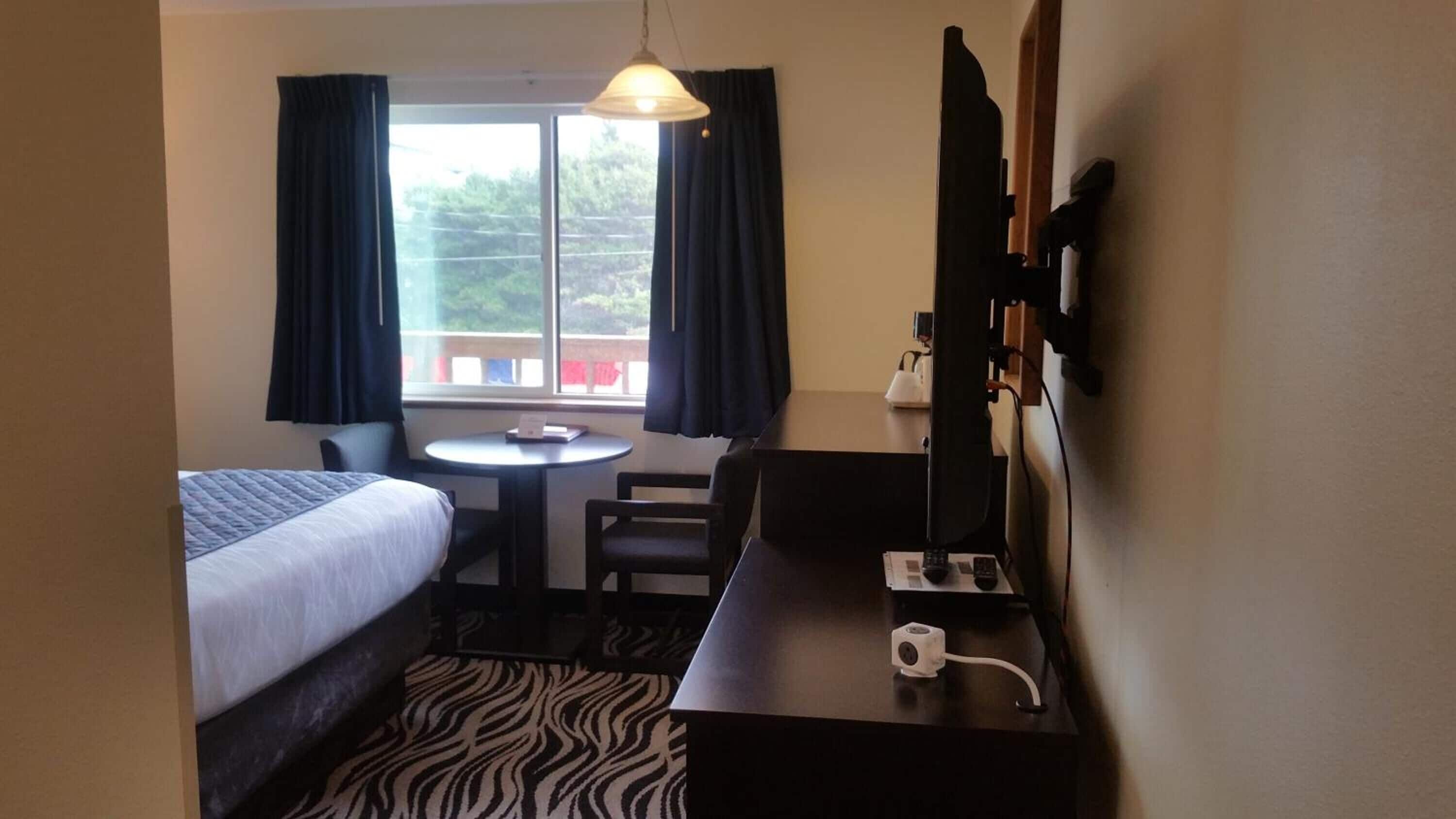 A hotel room with a bed, a desk with a computer, a chair, a small table, and a window with curtains.