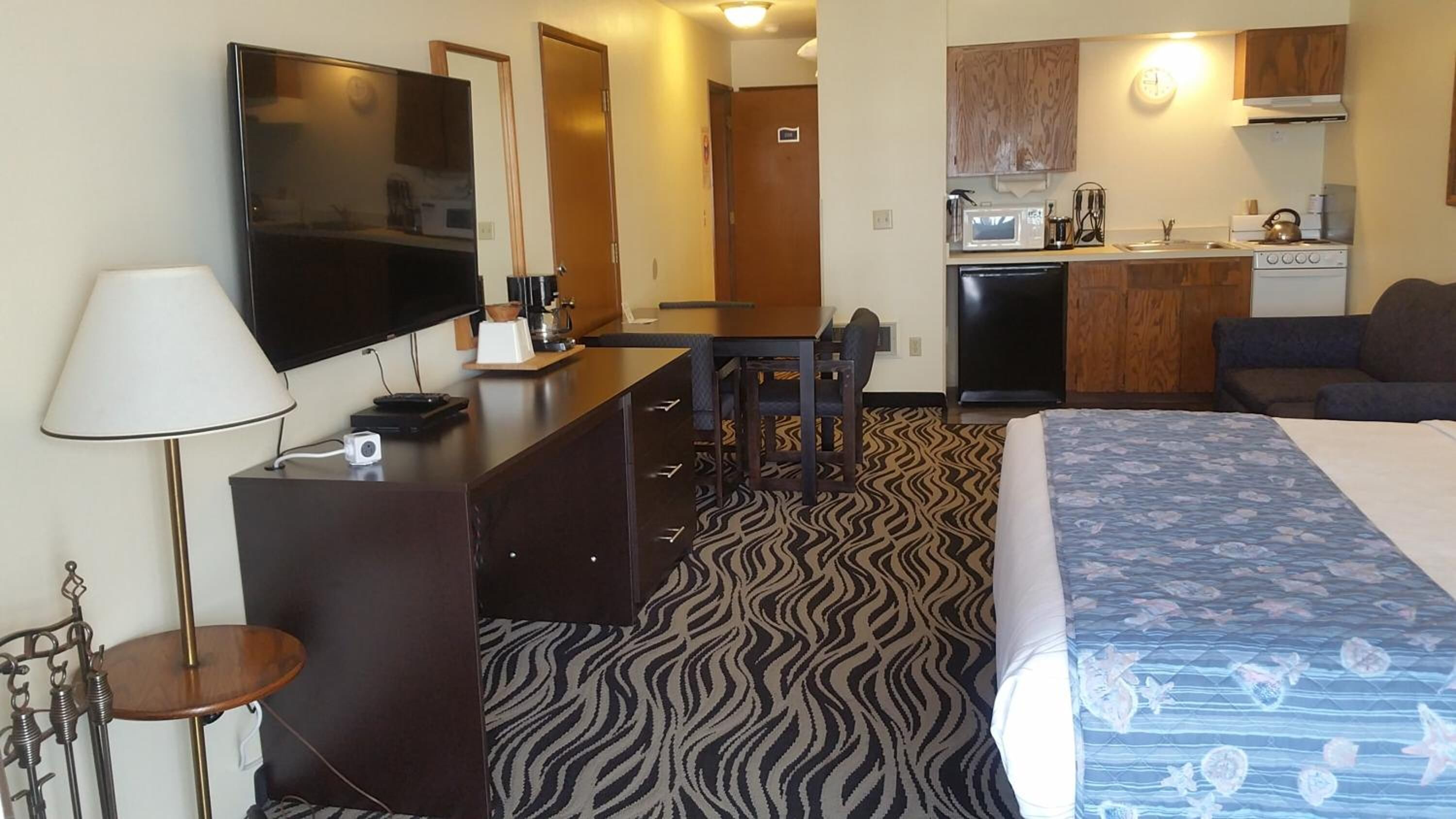 A hotel room with a bed, a desk with a flat-screen TV, a kitchenette, and a patterned carpet.