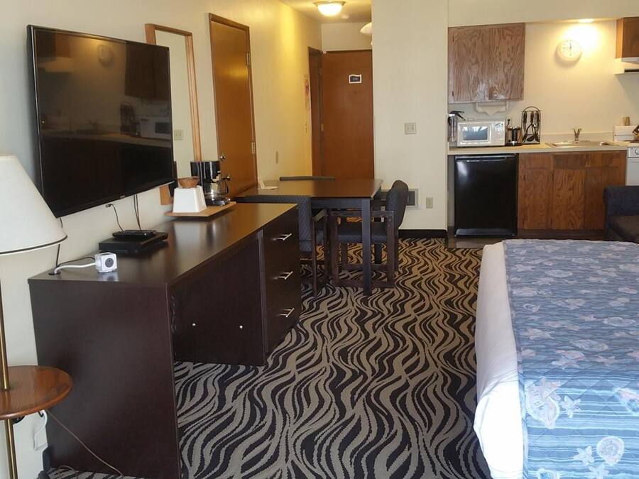 A hotel room with a bed, a desk with a flat-screen TV, a kitchenette, and a patterned carpet.