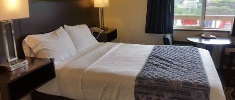 Quarto de hotel com cama, duas luminárias de cabeceira, telefone ao lado da cama, uma mesa pequena e uma janela com vista para o exterior.