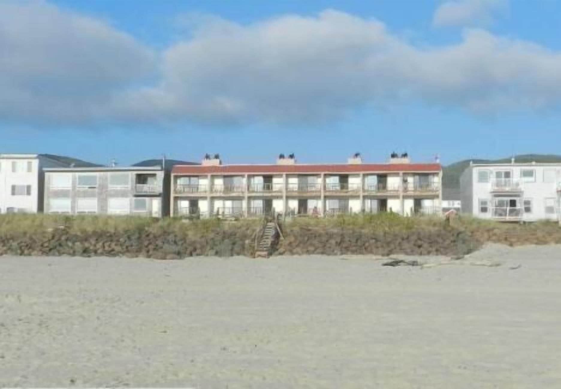 Un motel situé en bord de mer, avec plusieurs étages, des balcons et une plage de sable au premier plan.