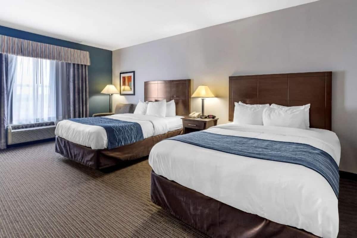 A hotel room with two beds, a large window with curtains, a wall-mounted lamp, and a framed picture.