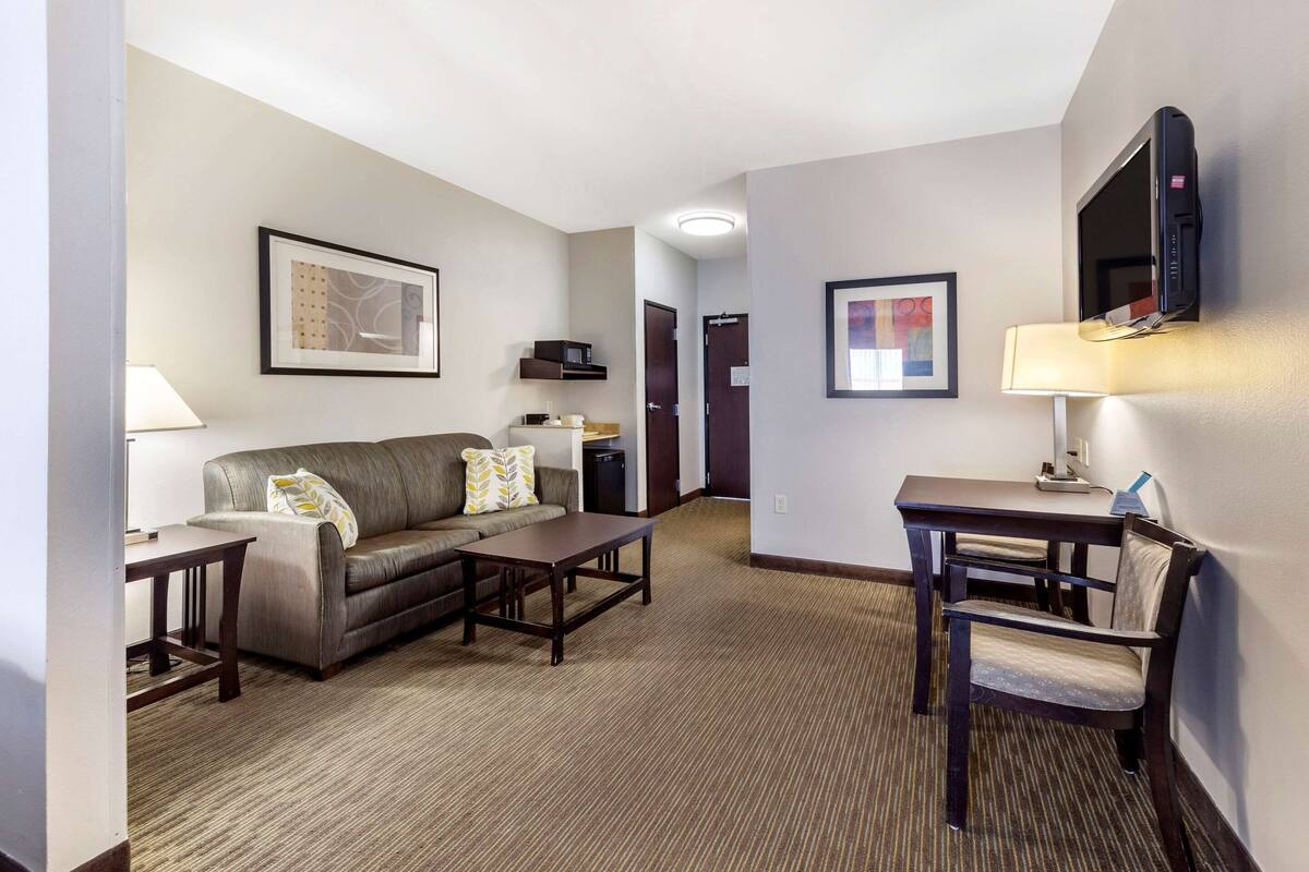 A hotel room with a sofa, a coffee table, a TV mounted on the wall, a desk with a chair, and framed artwork on the walls.