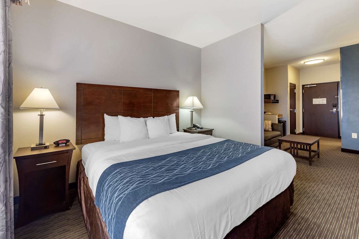 A hotel room with a large bed, two bedside tables with lamps, a small table, and a door leading to another room.
