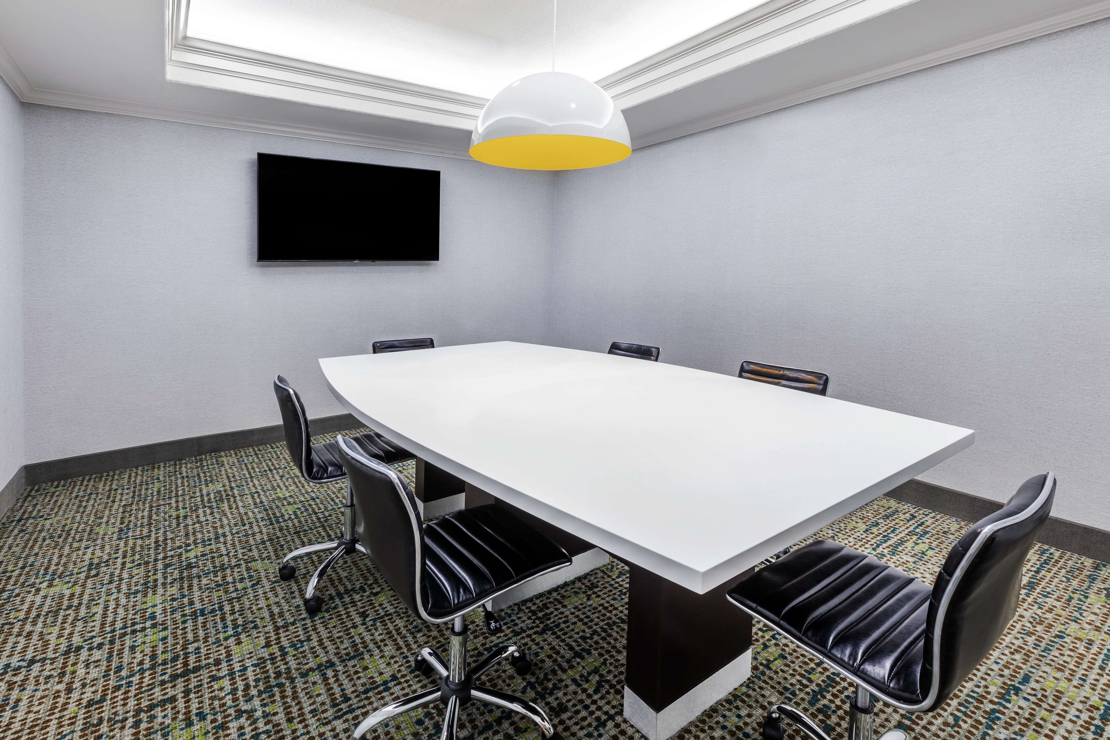 Une salle de conférence avec une table blanche, des chaises noires, un téléviseur fixé au mur et une moquette à motifs.