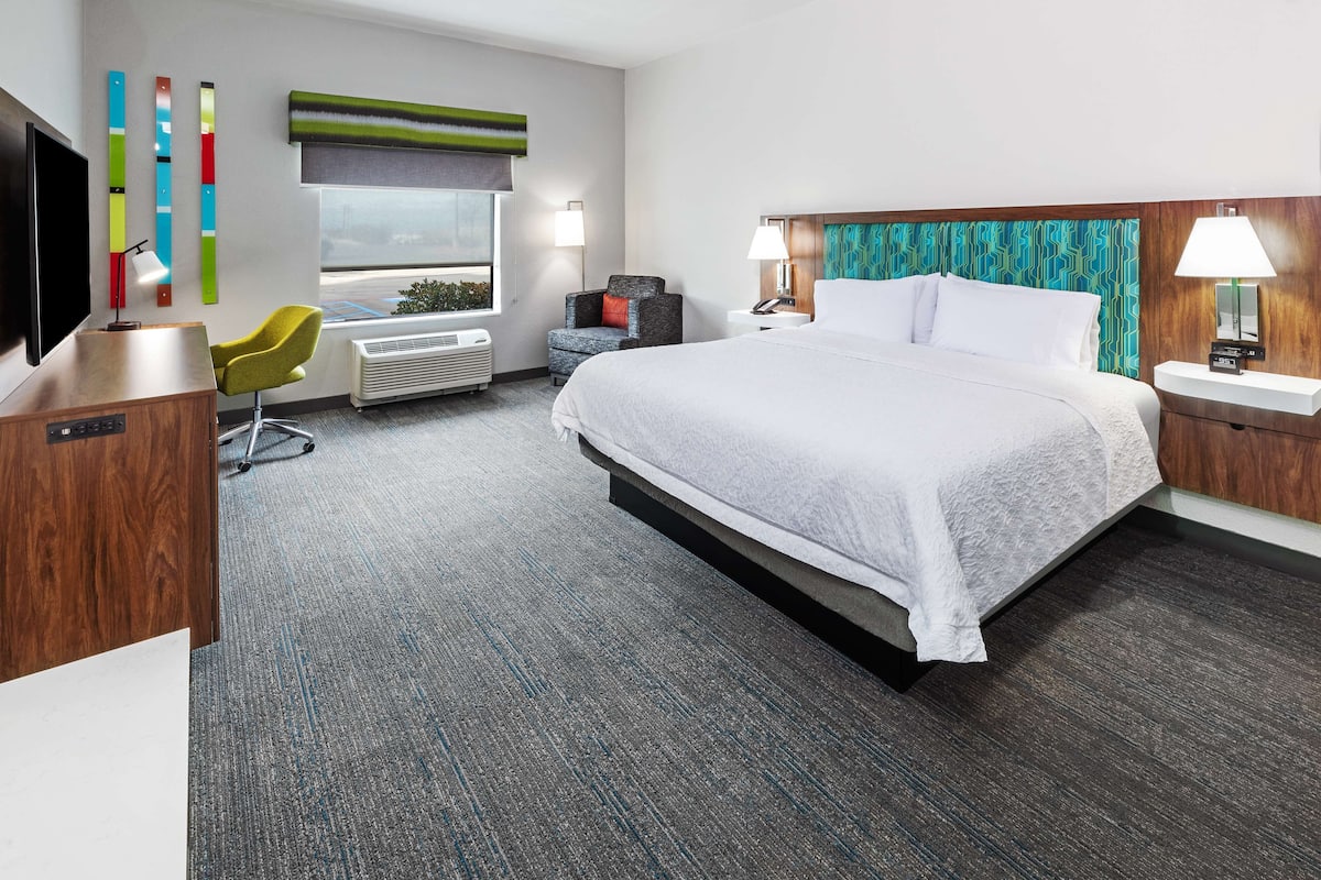 A hotel room with a large bed, a desk with a flat-screen TV, a chair, a desk lamp, and a window with a view.