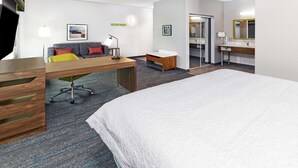 Une chambre d’hôtel avec un grand lit, un bureau avec une chaise, un canapé avec des coussins, une petite table, un miroir et une salle de bain avec un lavabo et un miroir.