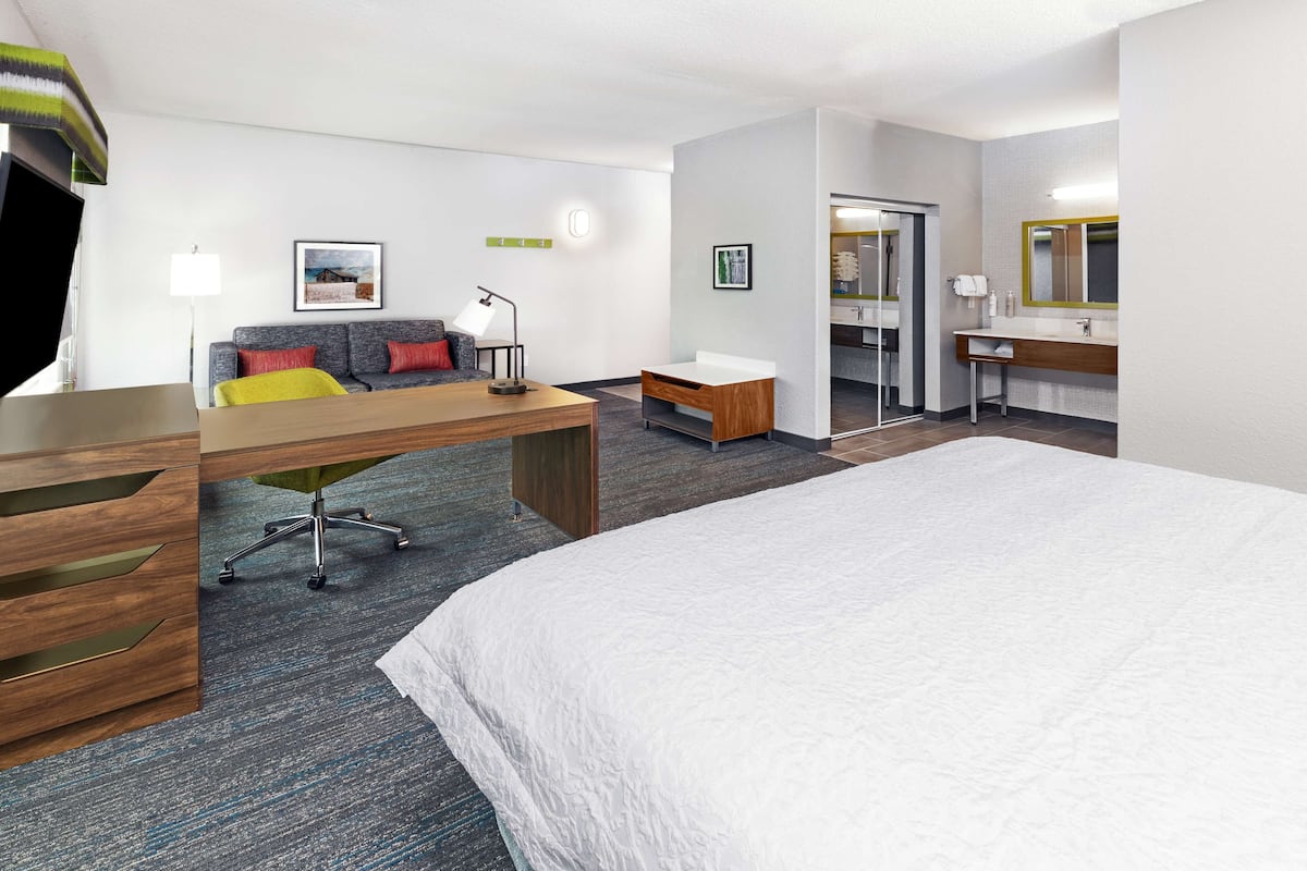A hotel room with a large bed, a desk with a chair, a sofa with cushions, a small table, a mirror, and a bathroom with a sink and mirror.