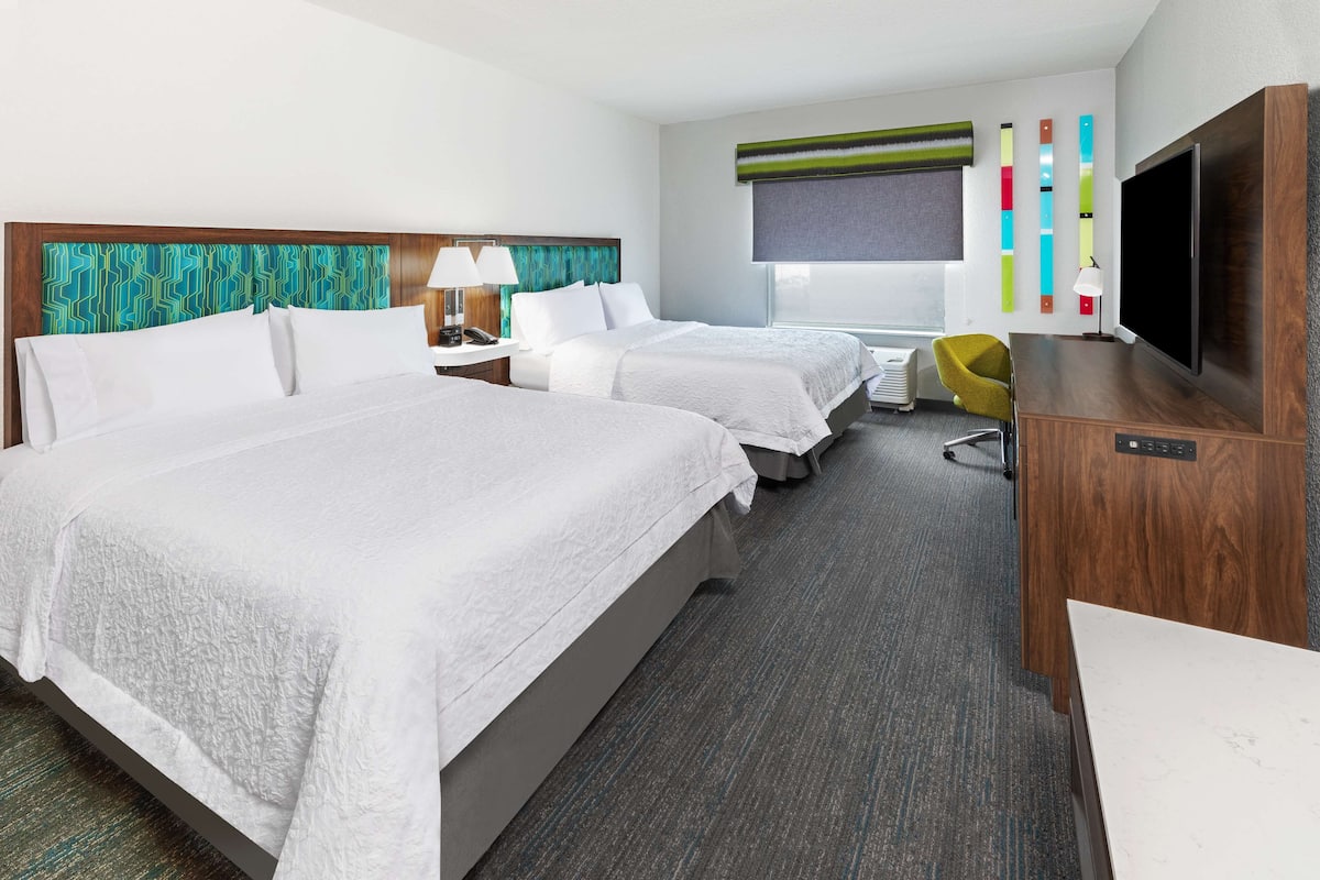 A hotel room with two beds, a desk, a TV, and a window with blinds.