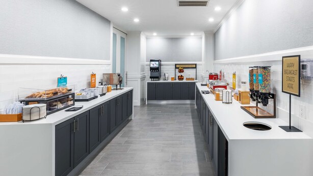 Un espace moderne dédié au petit-déjeuner, avec un long comptoir, des machines à café en libre-service et un choix varié de produits alimentaires.