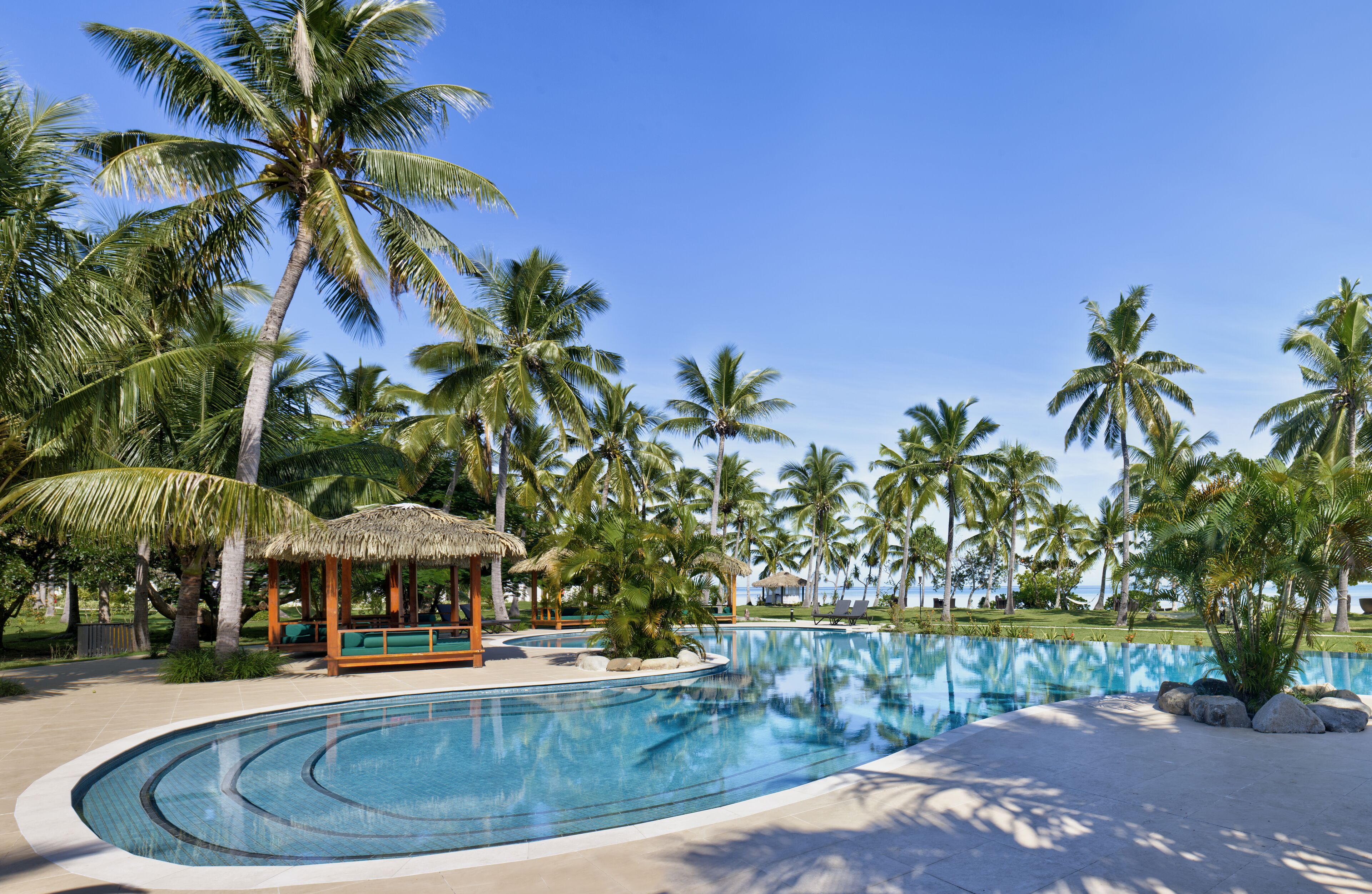 A resort pool area with palm trees, a thatched-roof gazebo, and a clear blue sky.