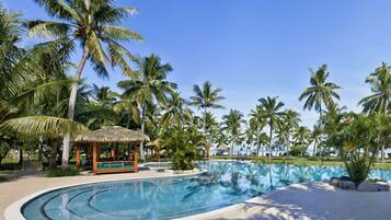 A resort pool area with palm trees, a thatched-roof gazebo, and a clear blue sky.
