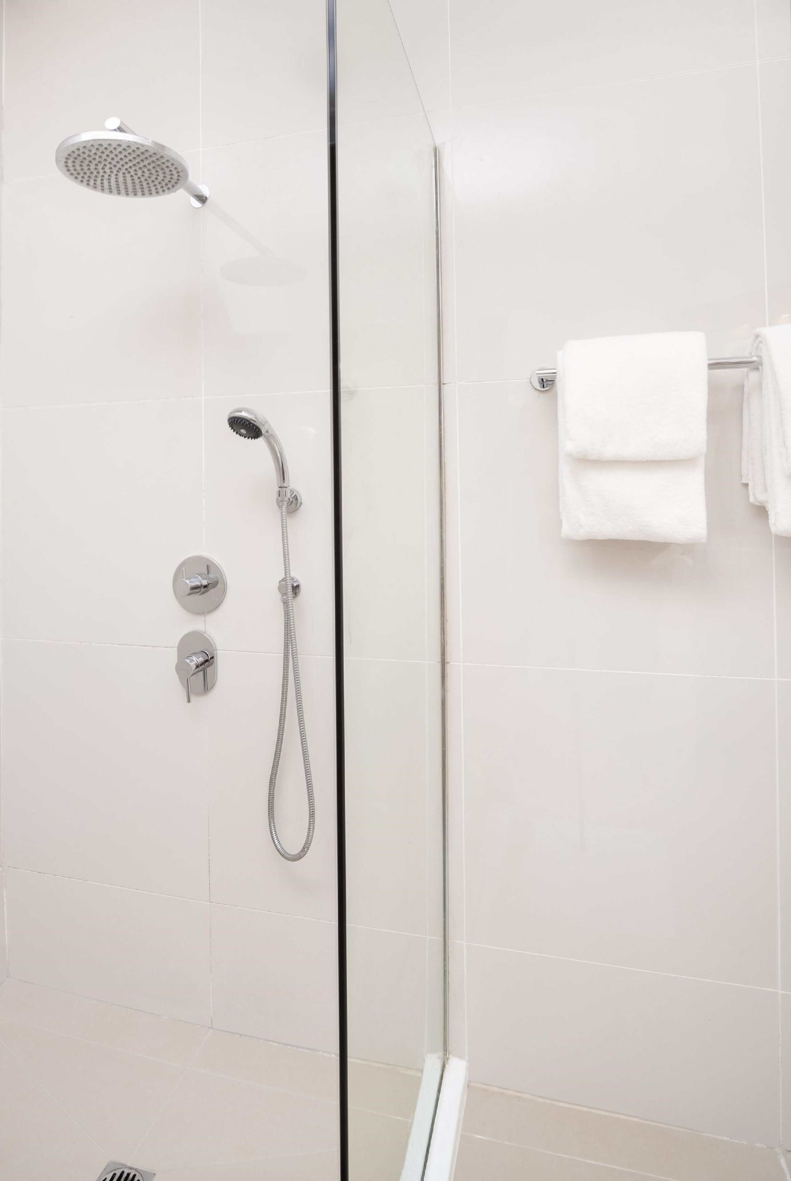 A modern shower with a handheld showerhead and a fixed showerhead, a glass door, and a towel rack with folded towels.