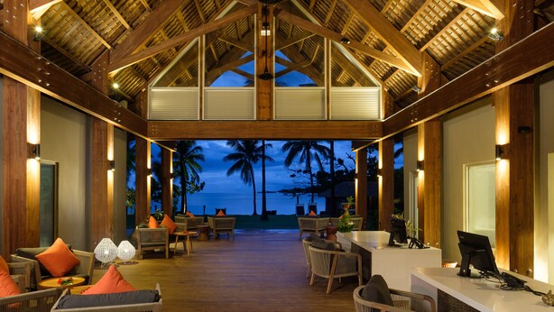 Un vaste espace de détente doté d’un plancher en bois et de hauts plafonds, comprenant plusieurs coins salon et offrant une vue sur la plage au crépuscule.
