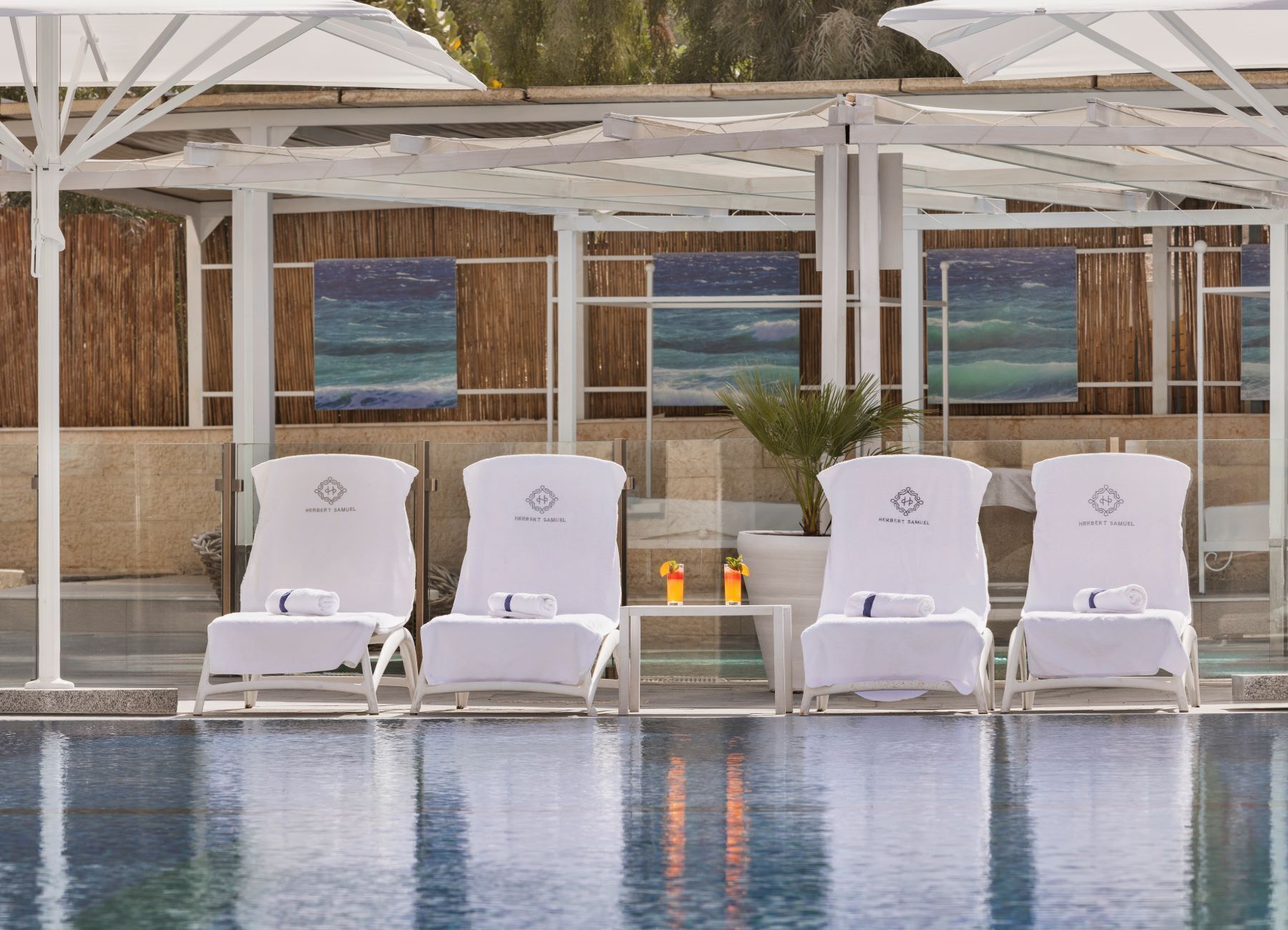 A poolside area with four white lounge chairs, each with a towel and a glass of a tropical drink. There are two large paintings of a beach scene on the wall behind the chairs.