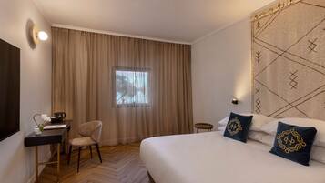 A hotel room with a bed, a desk with a chair, a window with curtains, and a wall-mounted lamp.