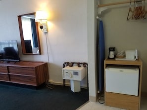 Habitación de hotel con tocador de madera, lavamanos pequeño blanco, espejo, lámpara, cortina de ducha y refrigerador miniatura.
