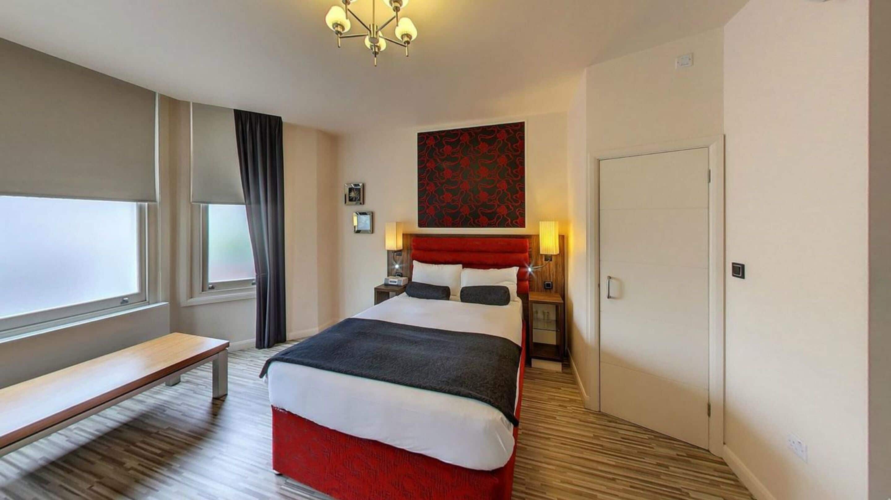 A hotel room with a bed, bedside tables, a bench, a window with curtains, a door, and a wall-mounted artwork.