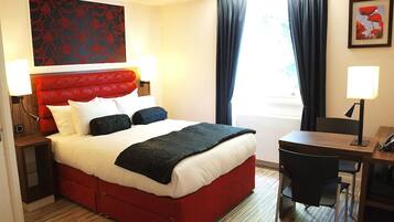 A hotel room with a red upholstered bed, white bedding, a desk with a chair, a lamp, and a framed picture on the wall.