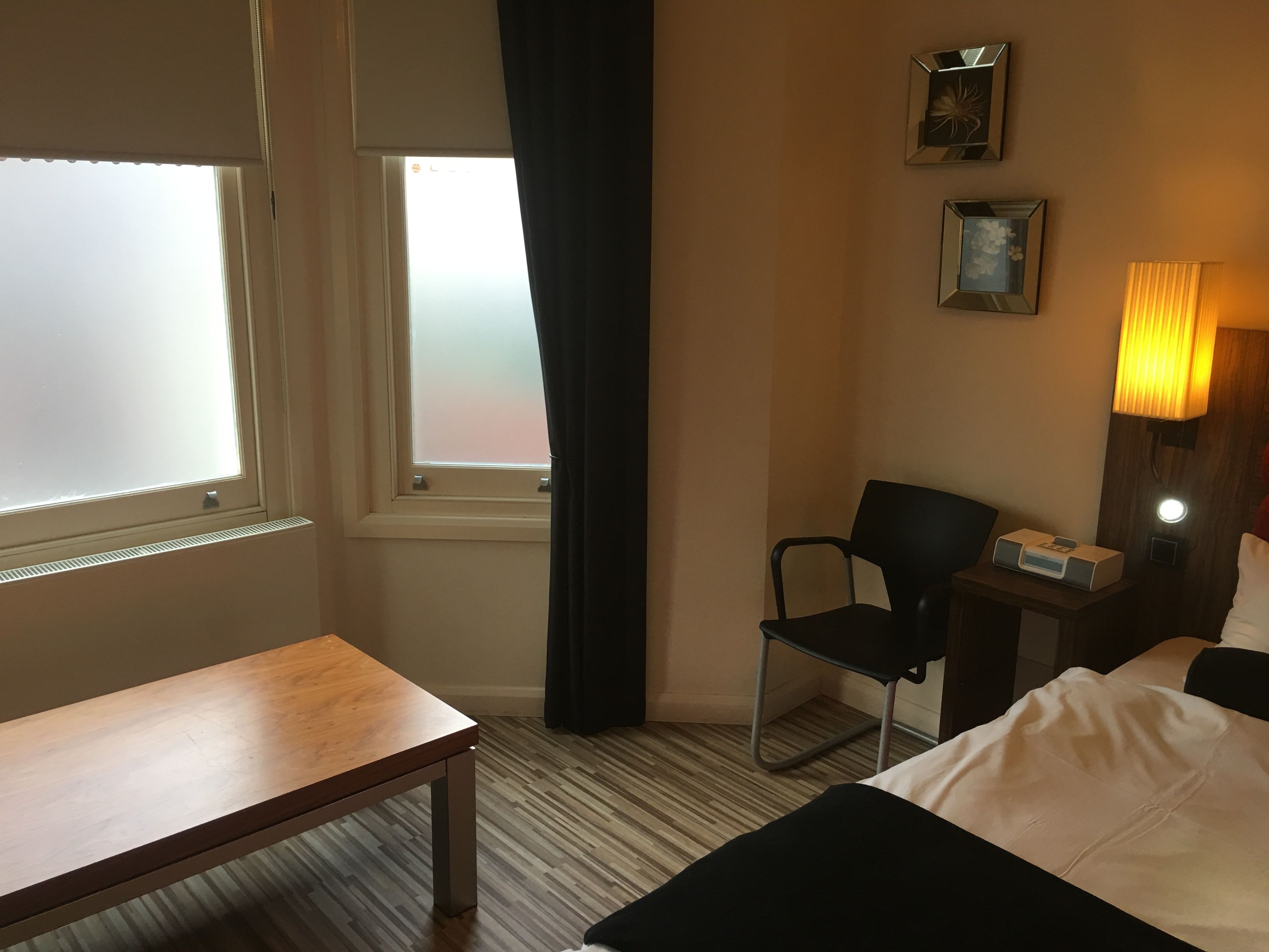 A hotel room with a bed, a desk, a chair, a nightstand, and a window with curtains.