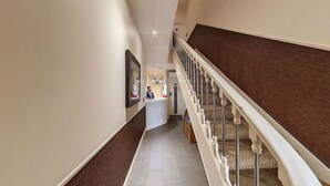A hotel reception area with a receptionist, a reception desk, and a staircase with a patterned carpet. - Simply Rooms & Suites (London)