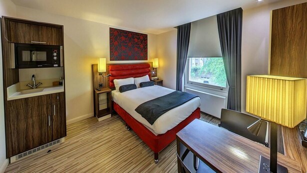A hotel room with a bed, a desk, a microwave, a sink, and a window with curtains. - Simply Rooms & Suites (London)