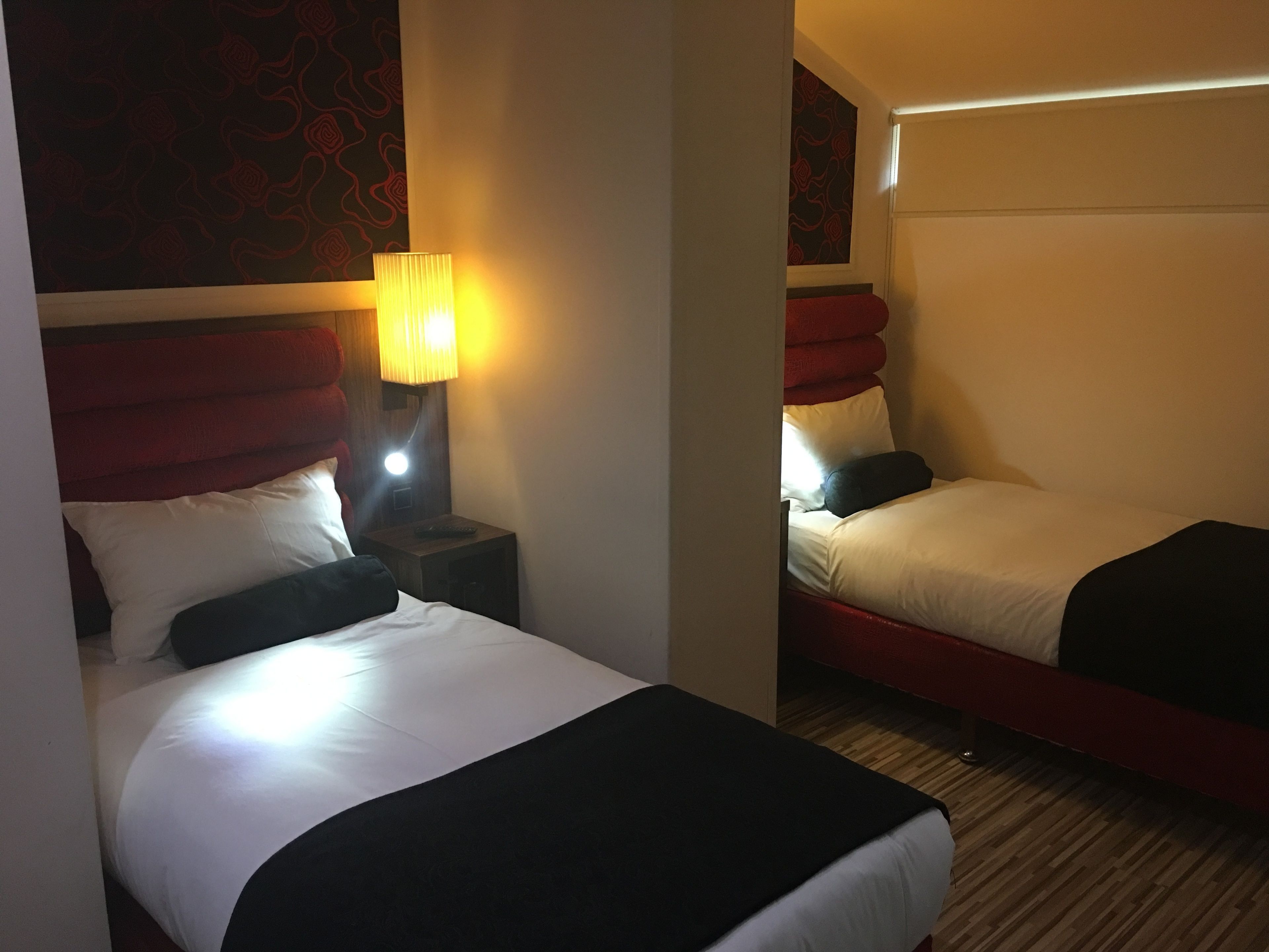 Una habitación de hotel con dos camas, una mesita de noche con lámpara y un cabecero rojo.