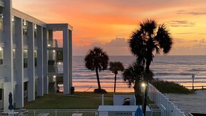 A pool area with lounge chairs and umbrellas, a building with balconies, and a view of the ocean at sunset. - Coastal Waters Inn (New Smyrna Beach)