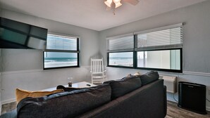A living room with a gray sofa, a white chair, a small table with a glass, and a ceiling fan. - Coastal Waters Inn (New Smyrna Beach)