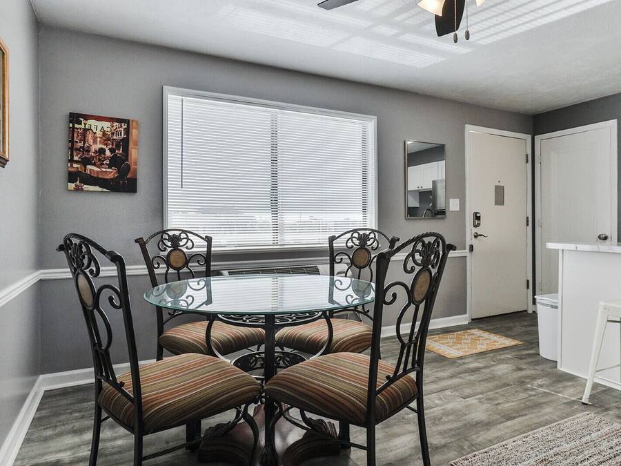 A dining area with a glass-top table and four chairs, a window with blinds, a framed picture on the wall, and a kitchen area with a white countertop and cabinets.