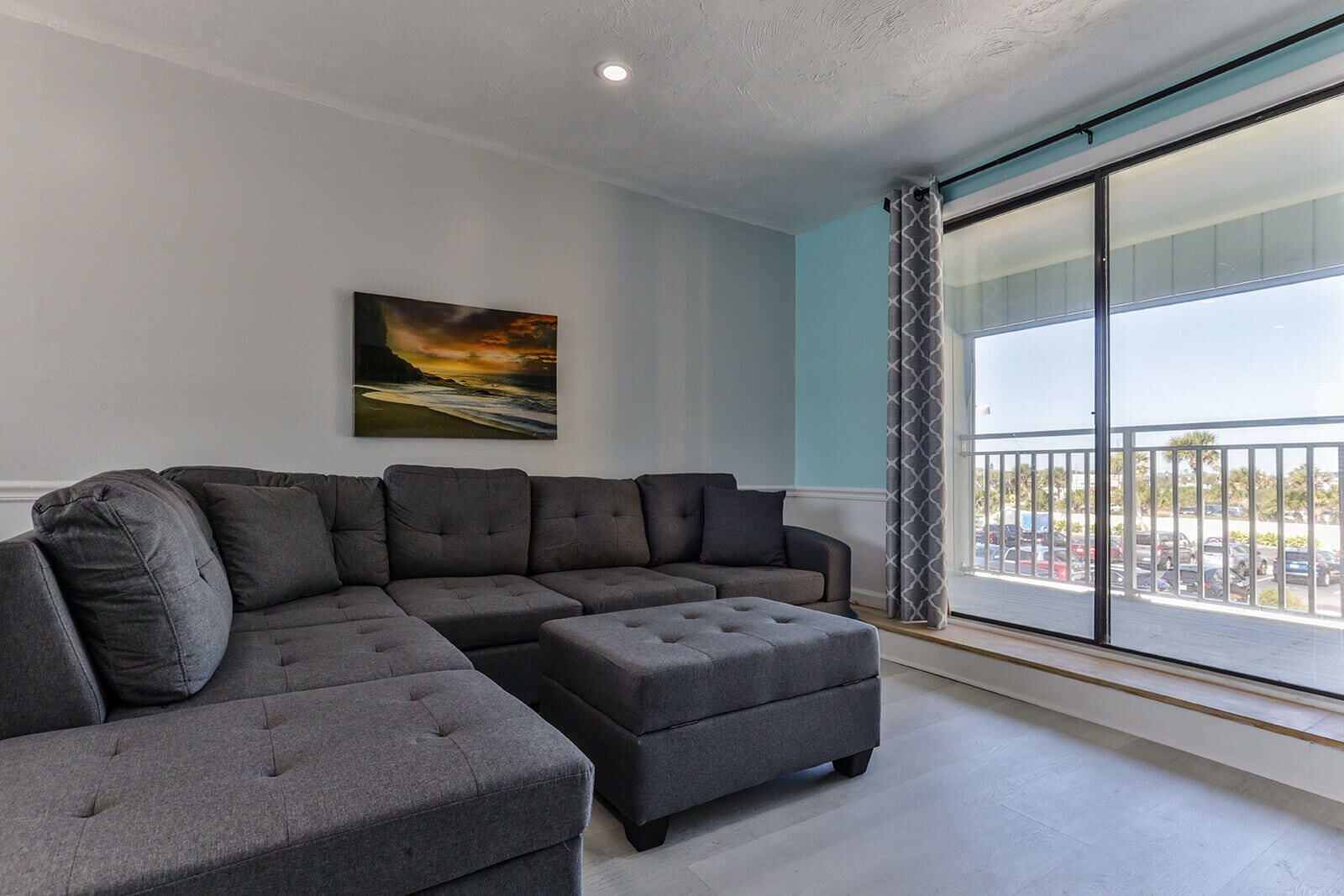 A modern living room with a grey sectional sofa and ottoman, a large window with a view of a parking lot, and a painting on the wall.