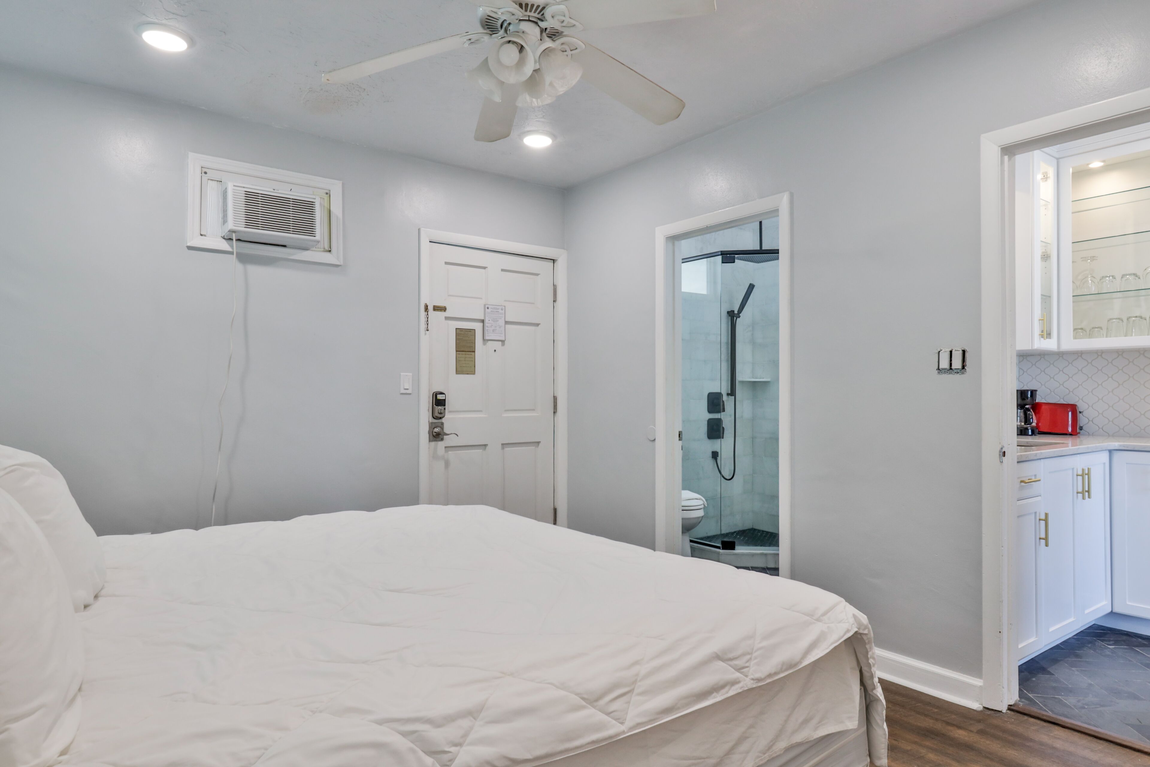 A bedroom with a bed, a ceiling fan, a white door, and a bathroom with a glass shower enclosure.