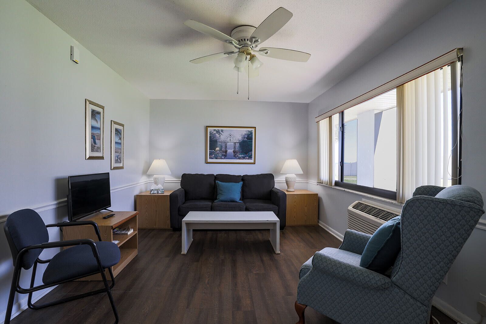A modern living room with a ceiling fan, a flat-screen TV, a sofa, a coffee table, and a large window with blinds.
