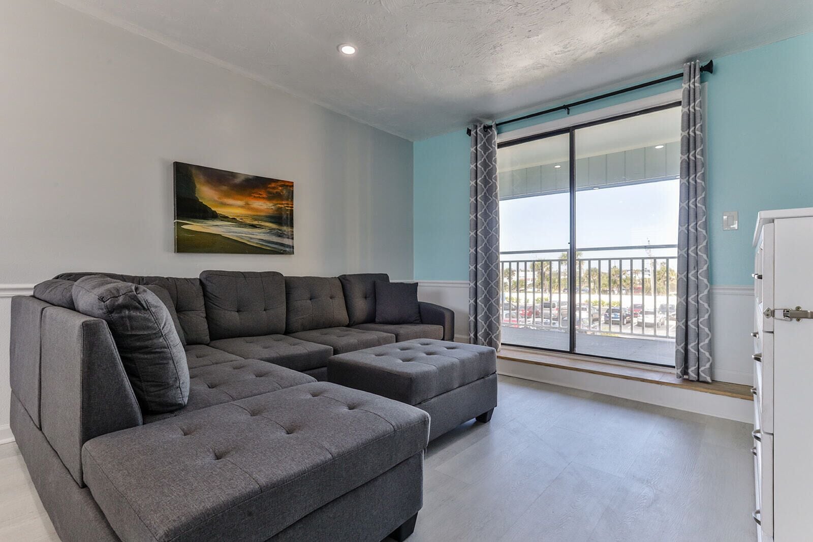 A modern living room with a sectional sofa, a painting, and a view of the outside through large windows.