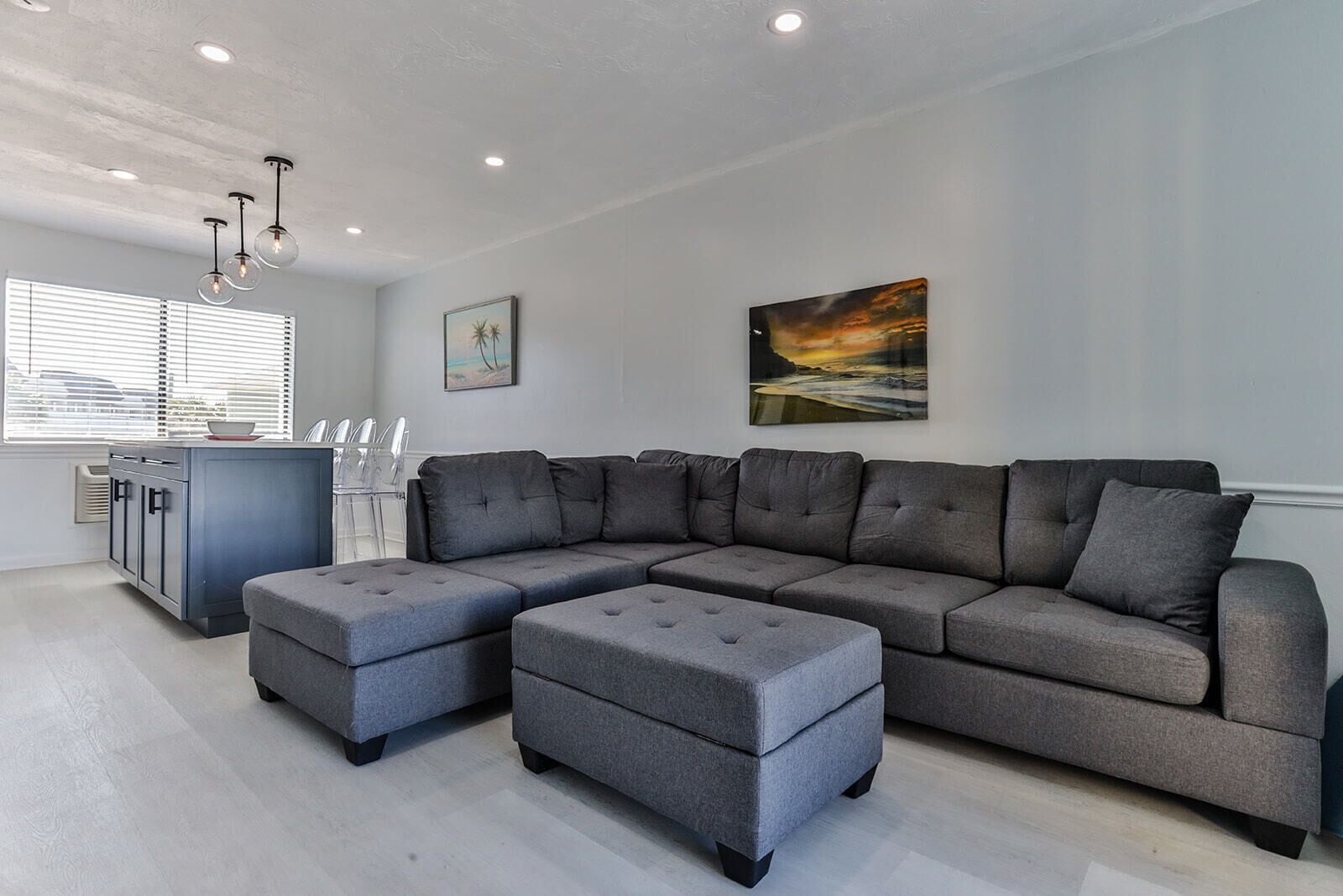 A modern living room with a grey sectional sofa, a grey ottoman, and a kitchen area with a fridge and bar stools.