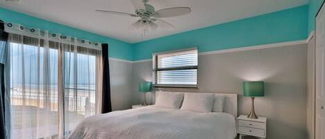 A bedroom with a bed, a ceiling fan, two bedside lamps, a window with blinds, and a view of the outdoors.