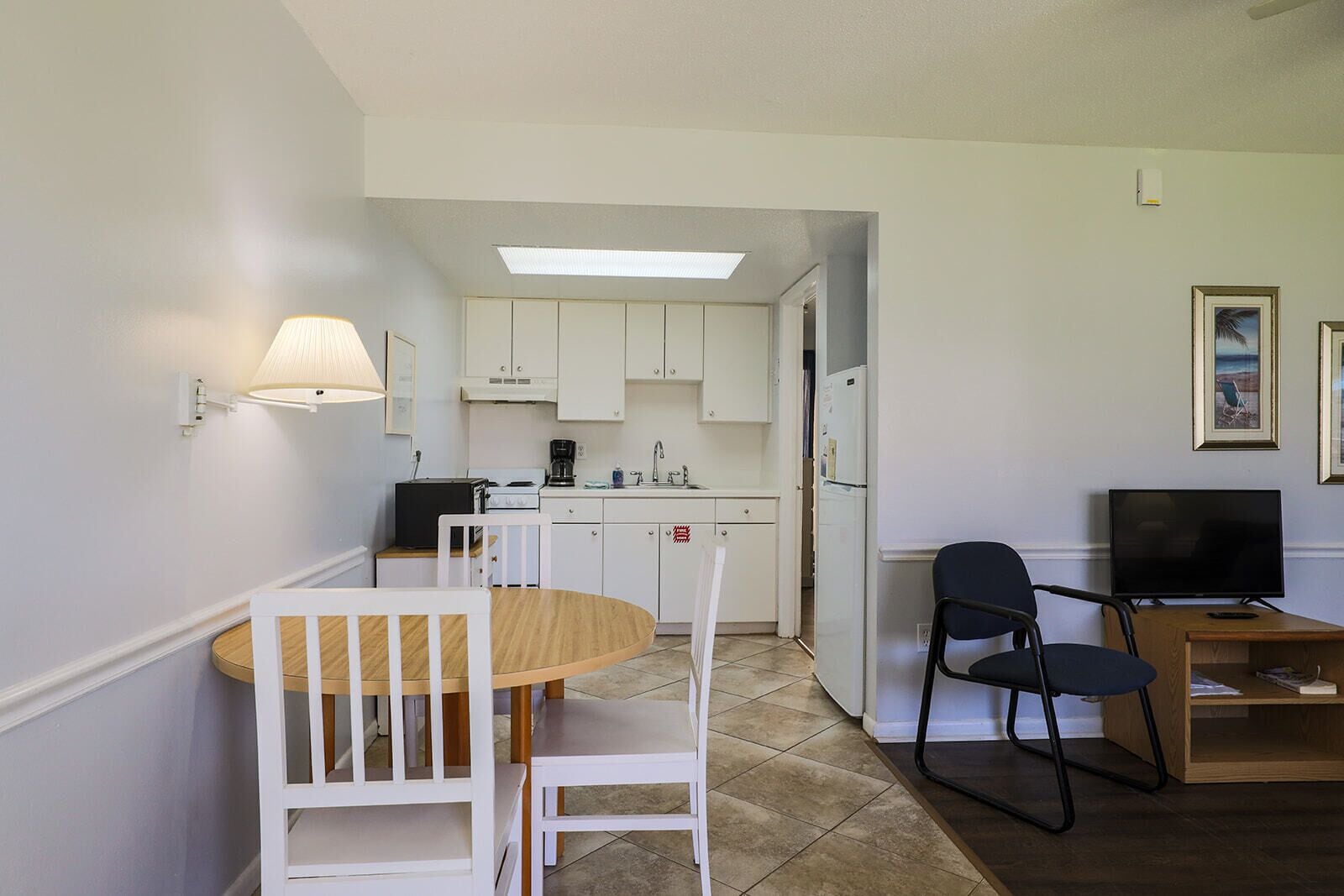 A compact kitchen with white cabinets, a dining area with a round table and chairs, a TV on a wooden stand, and a ceiling light.