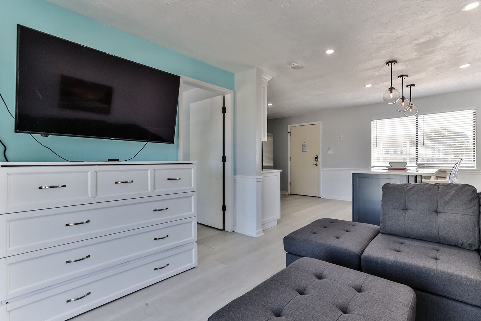 A modern living room with a flat-screen TV, white cabinetry, grey sofa, and a dining area with a window.