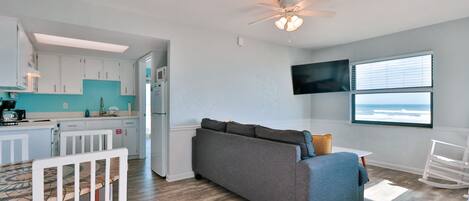 A modern living room with a gray sofa, a dining table with chairs, a ceiling fan, and a large window with a view.