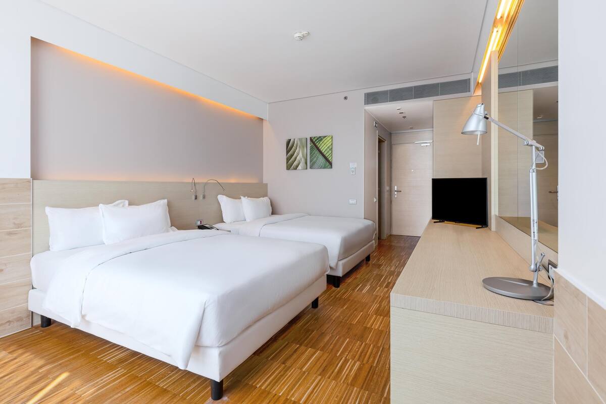 Habitación de hotel con dos camas, suelo de madera, mesita de noche con lámpara y un espejo grande.