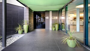 Entrada de un hotel con un corredor cubierto, plantas en macetas y una puerta de cristal que da al interior.