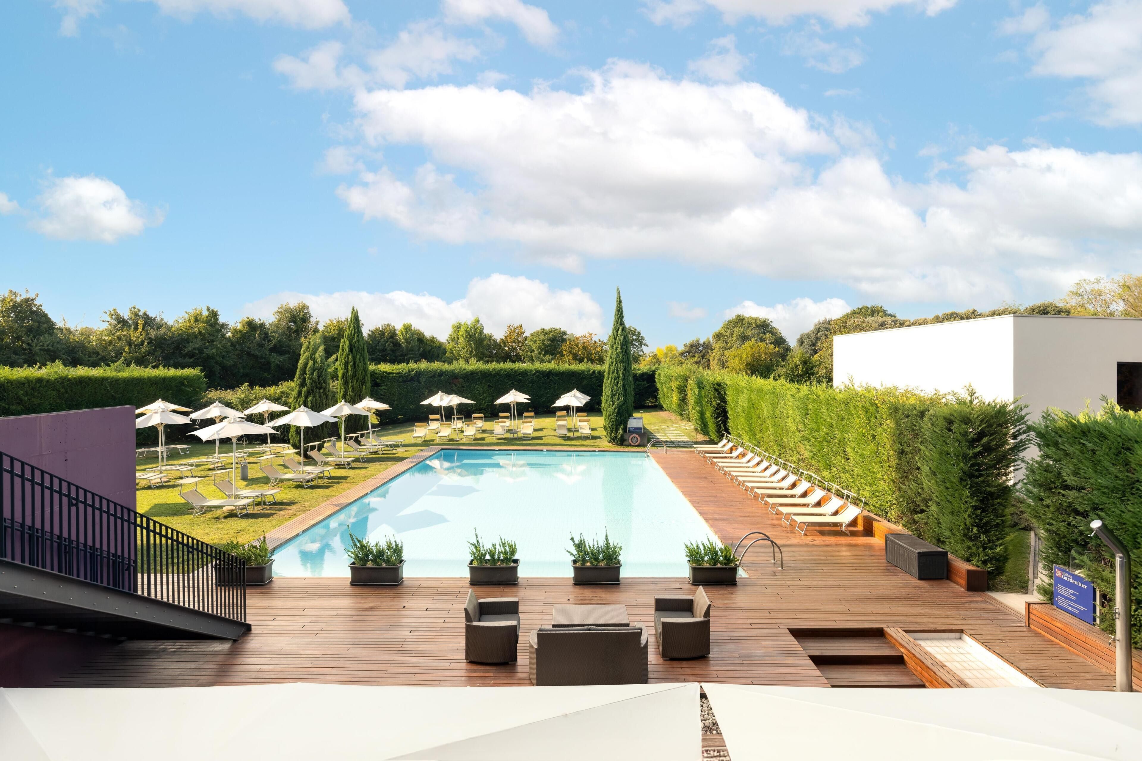 A swimming pool with lounge chairs and umbrellas, surrounded by greenery and a wooden deck.