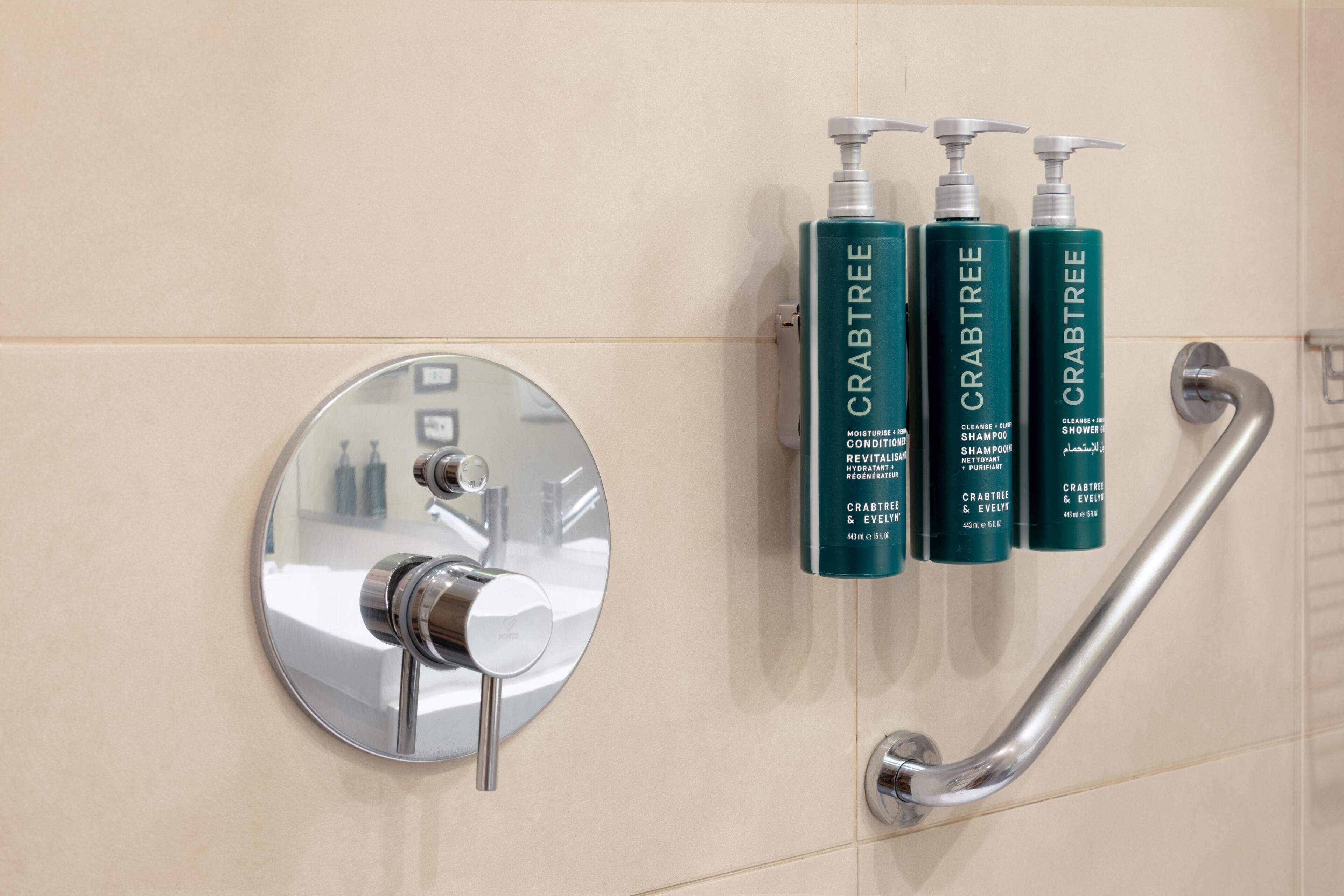 A bathroom with a modern sink, a handrail, and three bottles of Crabtree shower gel.