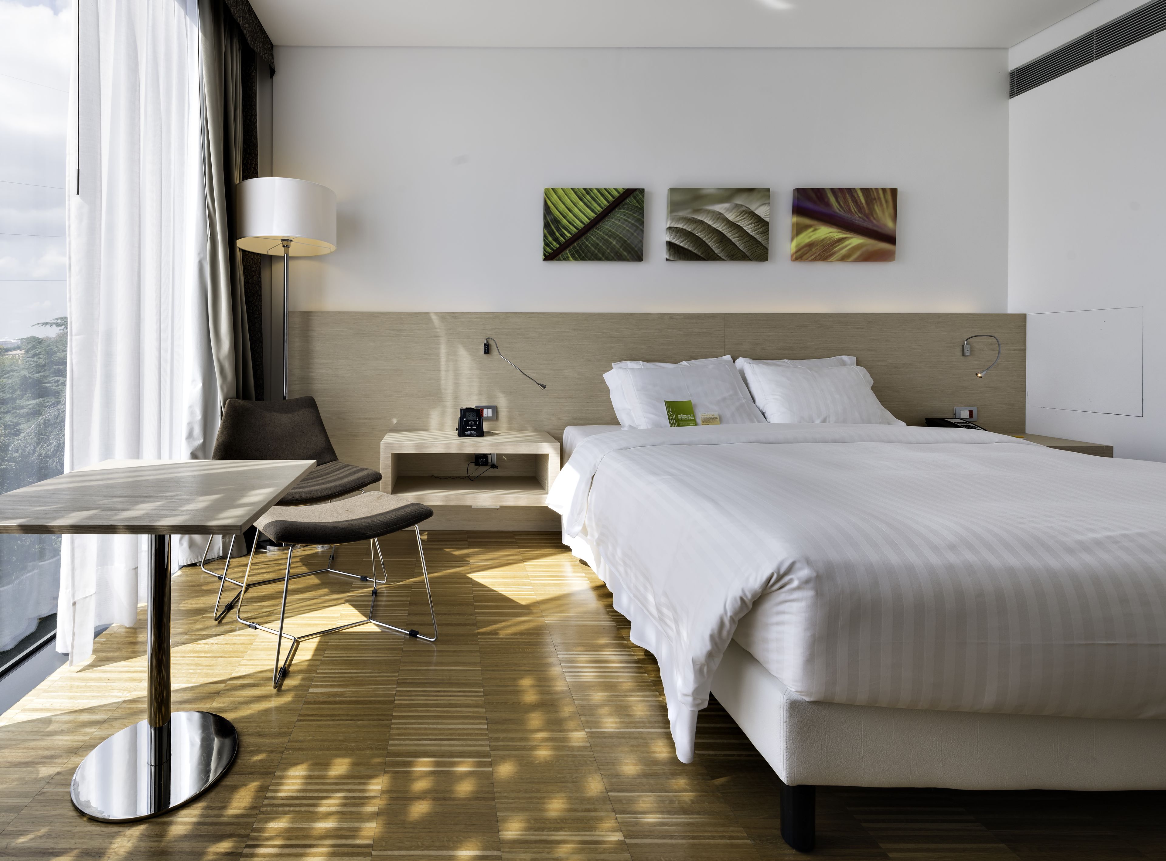A modern hotel room with a large bed, a desk with a chair, a bedside table, and a view of the outdoors.