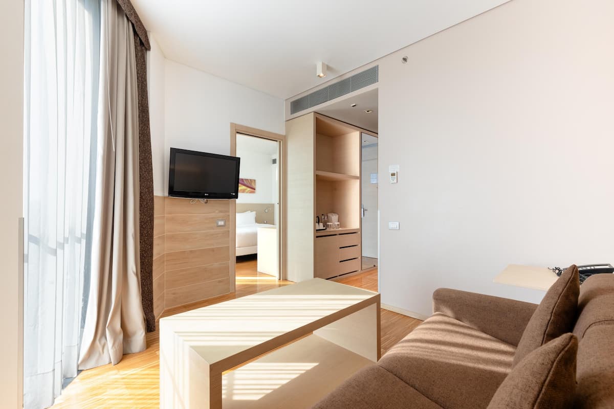 Una habitación de hotel moderna con sofá, televisor, piso de madera y una ventana grande con cortinas.