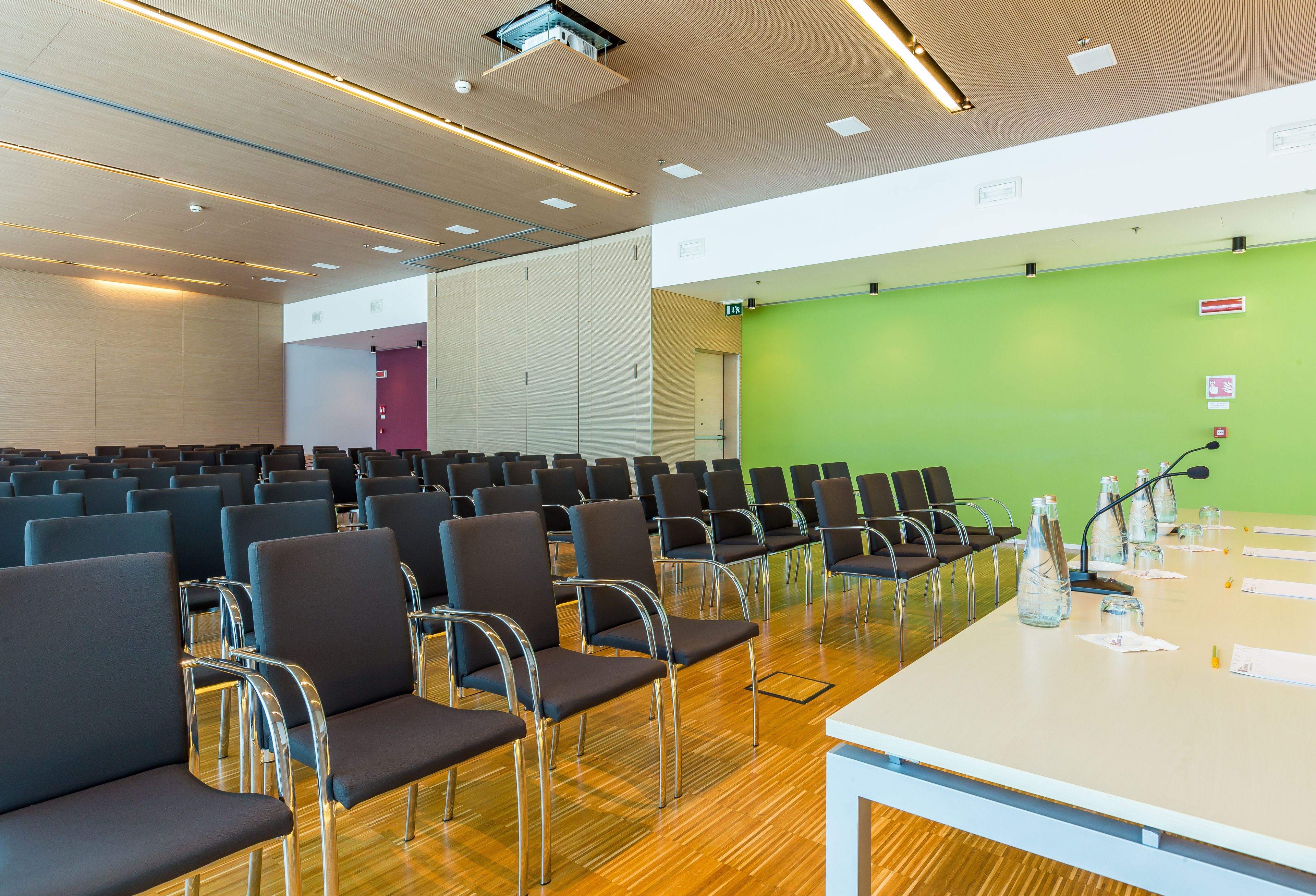 Ein Konferenzraum mit Reihen von Stühlen, einem langen Tisch mit Mikrofonen und einer grünen Leinwand.