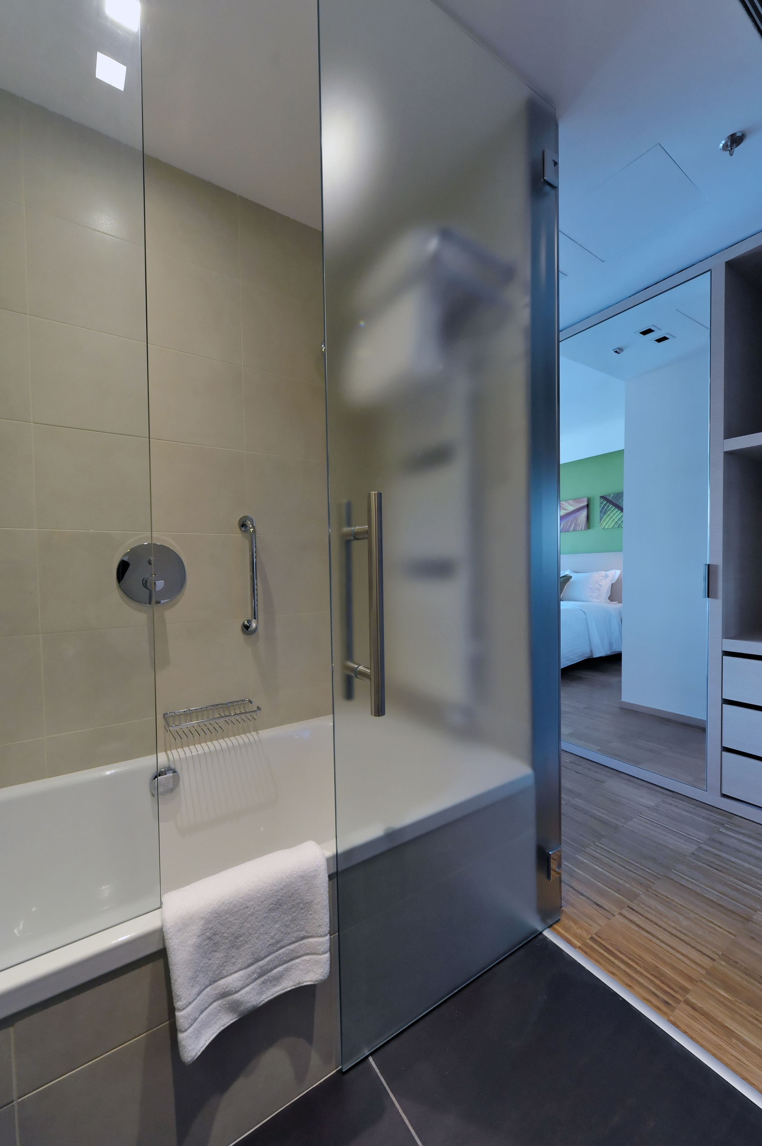 Un baño moderno con cabina de ducha de vidrio, una bañera blanca y una habitación visible con cama y paredes verdes.