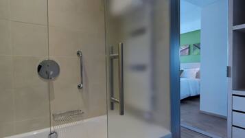 Un baño moderno con cabina de ducha de vidrio, una bañera blanca y una habitación visible con cama y paredes verdes.
