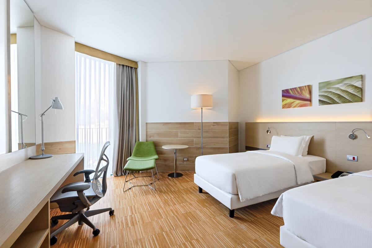 Una habitación de hotel con escritorio, dos camas, una silla, una lámpara y obras de arte en la pared.