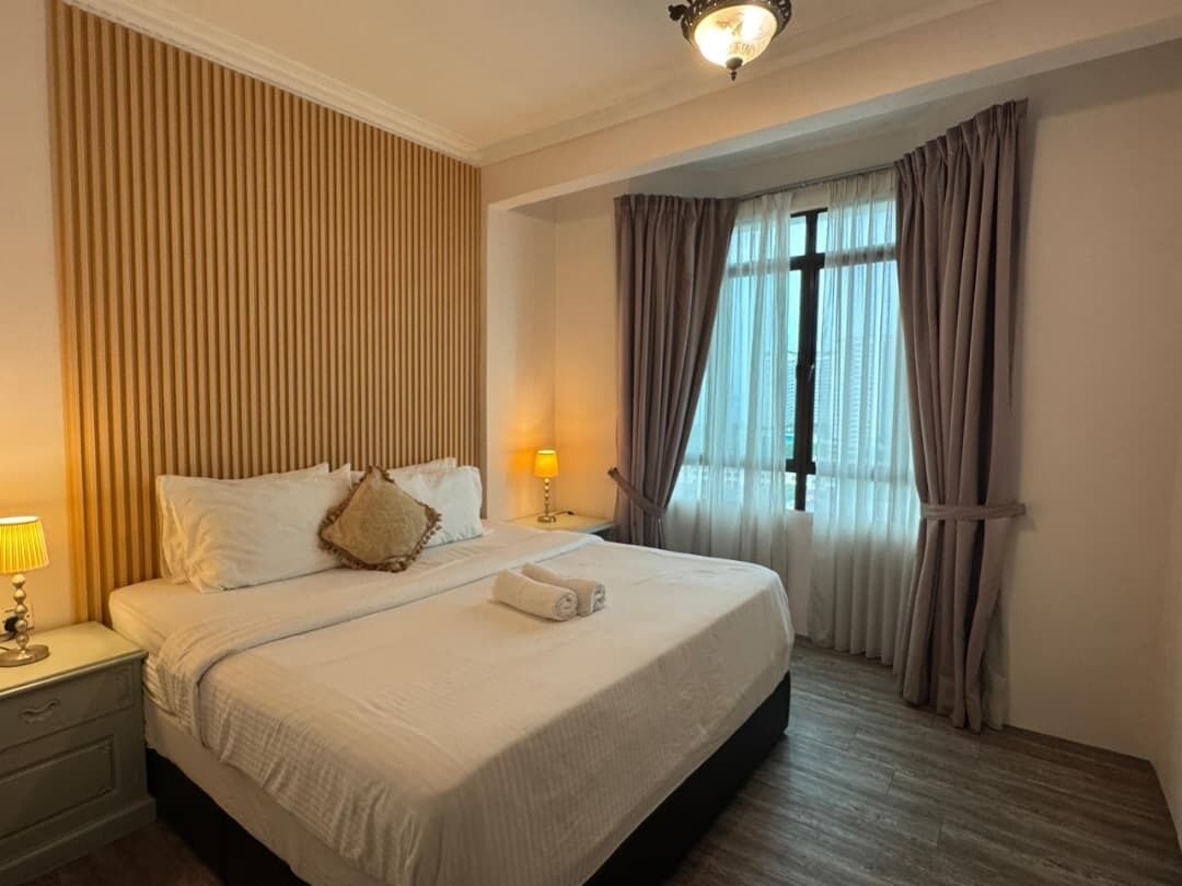 Una cama bien hecha con almohadas y una toalla doblada, una lámpara de noche y una ventana con cortinas.